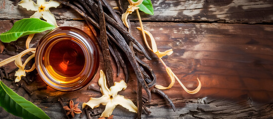 Vanilla extract in a glass bowl surrounded by vanilla pods, white blossoms, green leaves, star anise, and cloves on rustic wood.