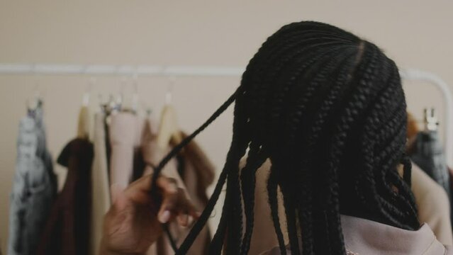 Back view slowmo shot of fashionable young Black woman with braided hairstyle choosing clothes on rack in minimalist apartment