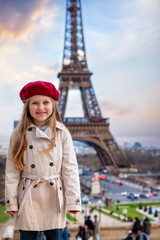 Portrait of a cute, blonde girl with a trench coat and a red beret hat in front of the Eiffel Tower in Paris, France