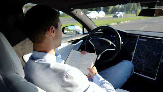 Male businessperson reading book during riding on electrical vehicle with autopilot at urban road. Successful businessman improving his knowledge while riding an autonomous self driving electric car