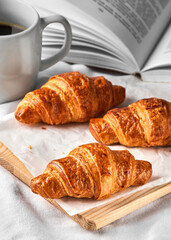 Fresh French croissants on white baking paper and wooden cutting board, a mug of coffee and opened book beside on a white bedding linen. Meal serve. Breakfast in bed, warm, pleasant and tasty morning 