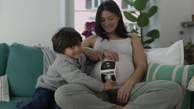 Happy Child Holding Ultrasound Image Beside Pregnant Mother - Genuine Family Bonding on Couch, Awaiting Unborn Baby Brother