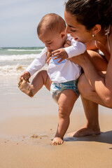 Vertical photograph of a young mother and her baby on the beach for the first time, baby touching the sand on her foot. concept family vacation with children on the beach in summer