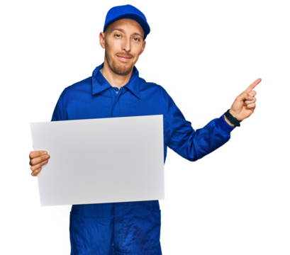 Bald man with beard wearing builder jumpsuit uniform holding empty banner smiling happy pointing with hand and finger to the side