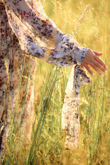 Woman in beautiful flower dress touching and waving high grass while walking in the meadow. Hand touch wild grass on the field. Close-up photo