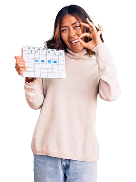 Young beautiful mixed race woman holding travel calendar doing ok sign with fingers, smiling friendly gesturing excellent symbol