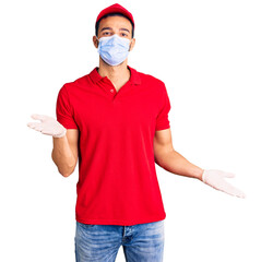 Young handsome hispanic man wearing delivery uniform and medical mask clueless and confused expression with arms and hands raised. doubt concept.