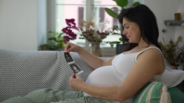 Happy Pregnant 30s woman seated on couch looking at ultrasound pictures of unborn baby, expecting child in third trimester pregnancy