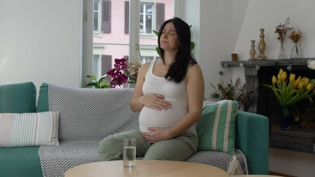 Pregnant woman having ache, 30s lady struggling with nausea during third trimester of pregnancy seated on couch sofa at home residence gently touching belly