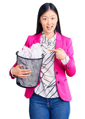 Young beautiful chinese woman holding paper bin full of crumpled papers smiling happy pointing with hand and finger