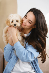 Brunette woman holding small maltipoo dog in her arms. Cute pet puppy friend at home