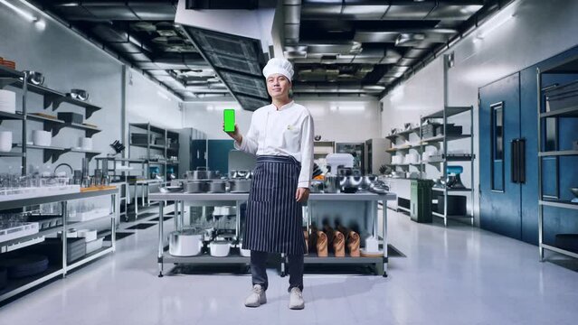 Full Body Of Asian Man Chef Smiling And Showing Green Screen Smartphone To The Camera While Standing In Kitchen