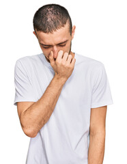 Hispanic young man wearing casual white t shirt smelling something stinky and disgusting, intolerable smell, holding breath with fingers on nose. bad smell