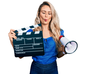 Young blonde woman holding video film clapboard and megaphone making fish face with mouth and squinting eyes, crazy and comical.