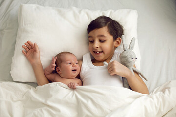 cheerful portrait of a boy of seven years old lying with his sister baby on a pillow on a white big bed with a crocheted toy hare , light from the window. - Powered by Adobe