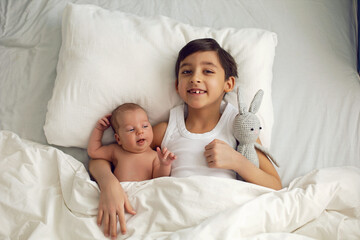 cheerful portrait of a boy of seven years old lying with his sister baby on a pillow on a white big bed with a crocheted toy hare , light from the window.