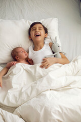 cheerful portrait of a boy of seven years old lying with his sister baby on a pillow on a white big bed with a crocheted toy hare , light from the window.