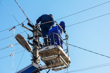 male electricians install new power lines on a pole at a height against a blue sky background - 767901208