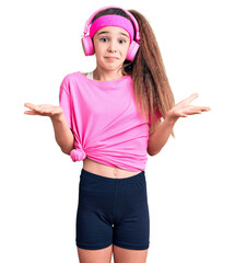 Obraz na płótnie Canvas Cute hispanic child girl wearing gym clothes and using headphones clueless and confused expression with arms and hands raised. doubt concept.