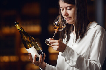 Professional woman sommelier smells white wine from glass, standing in cellar against shelf various alcoholic beverage background.