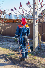 An electrician man in overalls and a red helmet stands at an electric pole and looks up