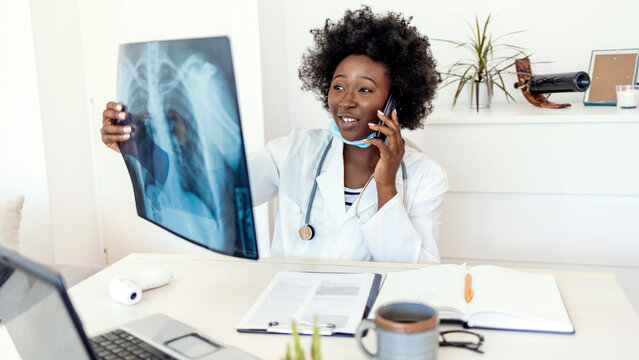 Healthcare, technology, roentgen, people and medicine concept. Female doctor in white coat with laptop computer looking at x-ray in medical office while talking via phone. Female doctor reviews x-ray.