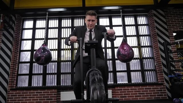 A successful, confident businessman in a black suit trains on a bicycle in a boxing gym. 