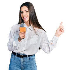Young brunette teenager holding reporter microphone smiling happy pointing with hand and finger to...