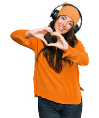 Beautiful brunette young woman listening to music using headphones smiling in love doing heart symbol shape with hands. romantic concept.