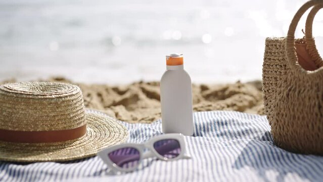Beach essentials arranged on blue striped towel. Sunscreen bottle, UV protection, alongside wicker accessories. Oceanfront scene depicts summer leisure, skincare necessity, vacation vibe. Slow motion.