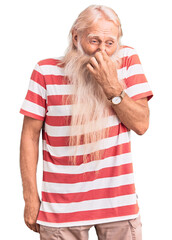 Old senior man with grey hair and long beard wearing striped tshirt smelling something stinky and...