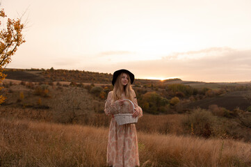 Young attractive woman in dress and hat sitting in grass on the hill at evening	