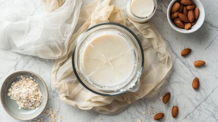 Top view of fresh homemade almond milk in a mason jar beside whole almonds and oats, on a marble surface with a muslin cloth.
