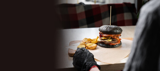 View over the shoulder of a woman in black gloves about to eat a delicious hamburger sitting at a table in a restaurant. Fast food lunch concept