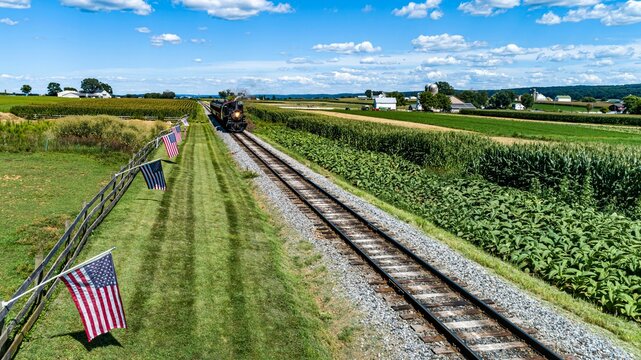 Aerial View of a Steam Passenger Train Approaching, on a Single Track With a Fence and America Flags