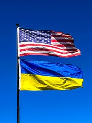American and Ukrainian flags waving in the wind against a bright blue sky mounted on a metal pole.