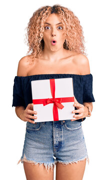 Young blonde woman with curly hair holding gift scared and amazed with open mouth for surprise, disbelief face