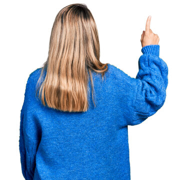 Young blonde woman wearing casual sweater posing backwards pointing ahead with finger hand