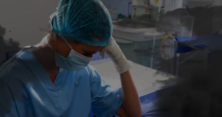 Image of black clouds over sad biracial female nurse with face mask in hospital