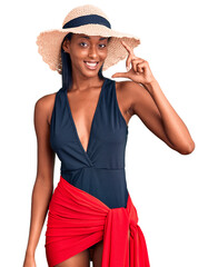 Young african american woman wearing swimsuit and summer hat smiling and confident gesturing with hand doing small size sign with fingers looking and the camera. measure concept.