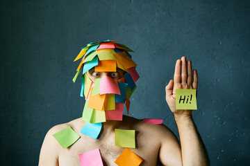 Funny man with cross-eyed covered with colorful sticky notes all over his face and head waving hello - 767959887
