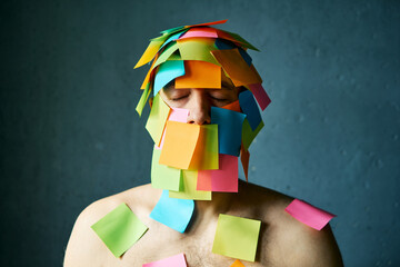 Close up portrait of man with closed eyes and colorful sticky notes all over his face and body over gray background - 767960010