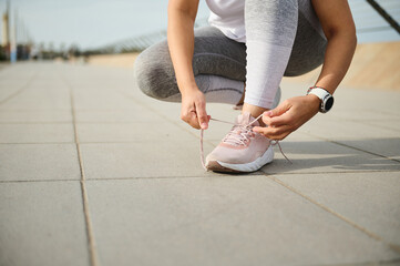 Close-up of a woman athlete runner jogger in gray leggings, tying laces on her pink sneakers,...