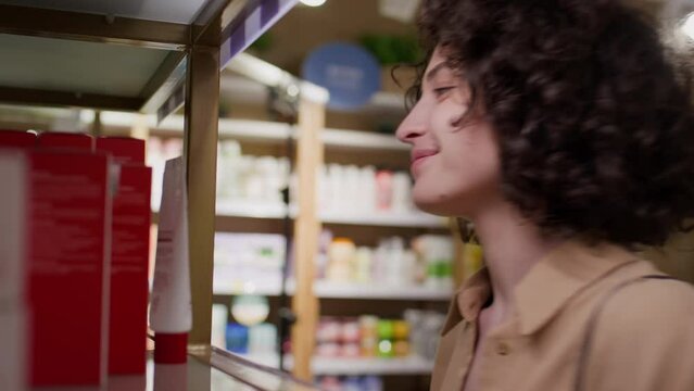 Medium close-up shot of young Caucasian woman with curly auburn hair picking up jar of cream from shelf in cosmetics store, looking at label, smiling, putting it into basket and walking away
