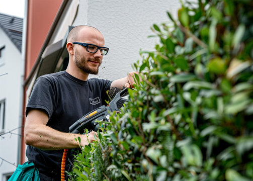 product photography of a man using powerful and imposing garden power tool, a cordless hedge trimmer with a long blade to cut the top of green hedges in a mid size modern house garden