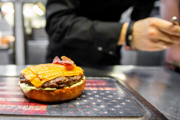 Freshly prepared cheeseburger with bacon on a star-patterned surface in a bustling kitchen,...