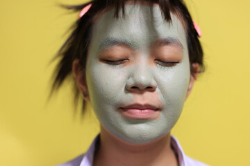 Lifestyle of a 14-16-year-old teenage girl applying a green clay facial mask. The teenager is doing an organic anti-blemish skincare routine at home.