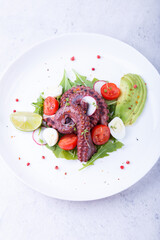 Warm salad with octopus, cherry tomatoes, avocado, arugula, quail eggs, radish and lime on a white plate. Traditional Mediterranean dish. Close-up, white background.