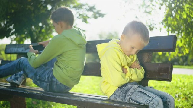 Young boy in bright yellow hoodie sitting on park bench, sulking with arms crossed while his brother absorbed in using smartphone. Children engage with world and each other