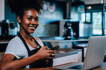 Close up portrait of concentrated black woman coffee shop owner in apron using smartphone in her...
